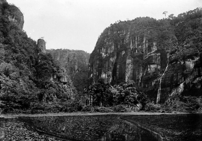 A photograph of a narrow gorge: sheer rock walls rising on both sides, thick forest at their feet, a thin waterfall dropping down the right-hand cliff and still water across the foreground.