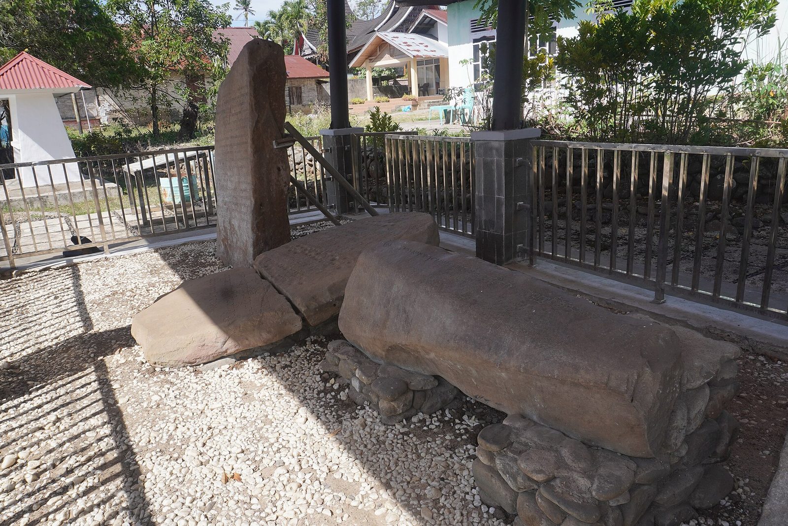 Several weathered stone slabs lying and standing on gravel inside a railed enclosure under a roof, their surfaces worn and their lettering barely legible, with houses and trees beyond.