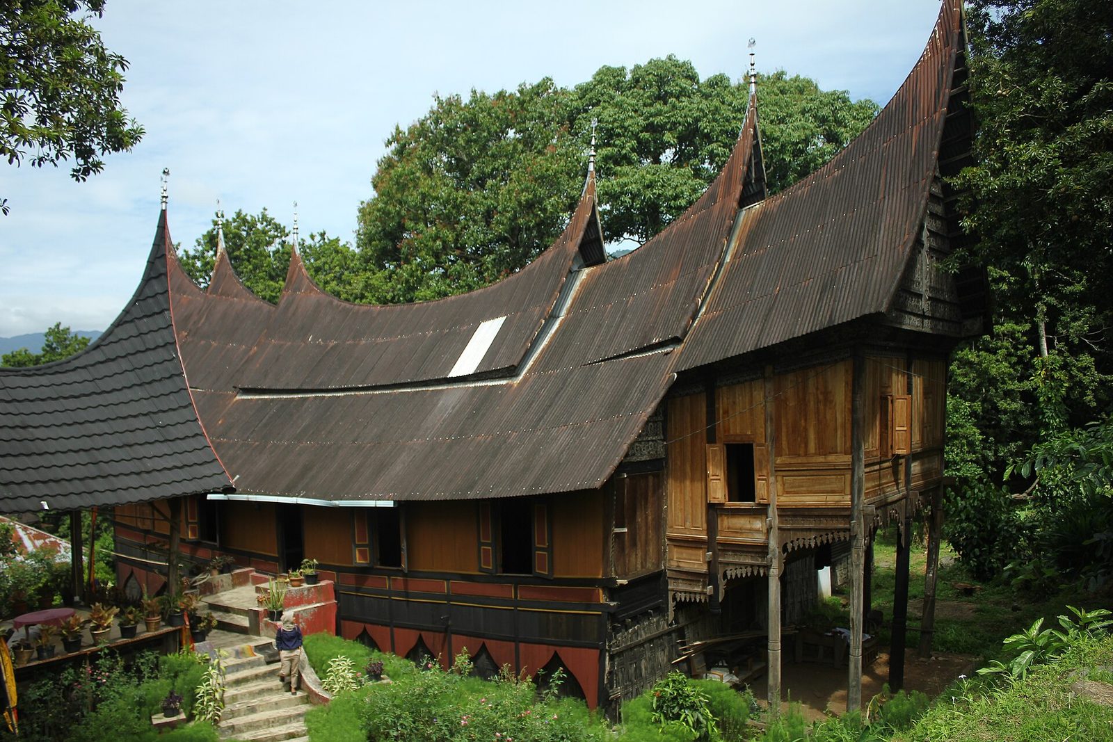 A long timber house raised on posts, its dark roof sweeping up into pointed gables at both ends, a carved and painted front wall below, and forest rising close behind it.