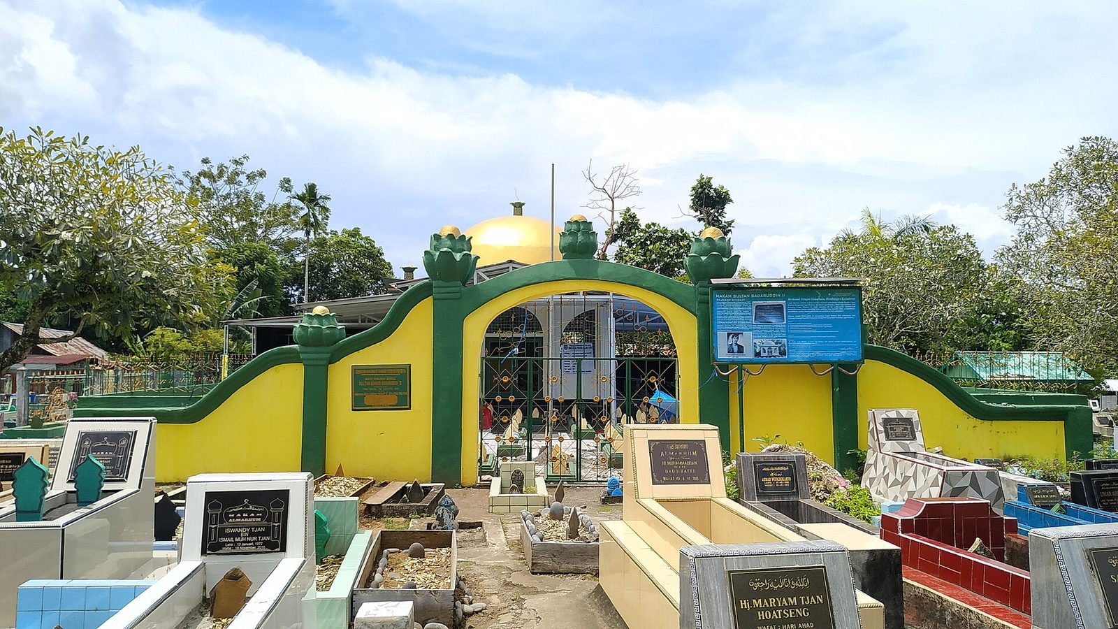 A walled cemetery entered through a green and yellow painted arch, rows of tiled and kerbed graves with inscribed headboards inside it, and trees along the boundary.