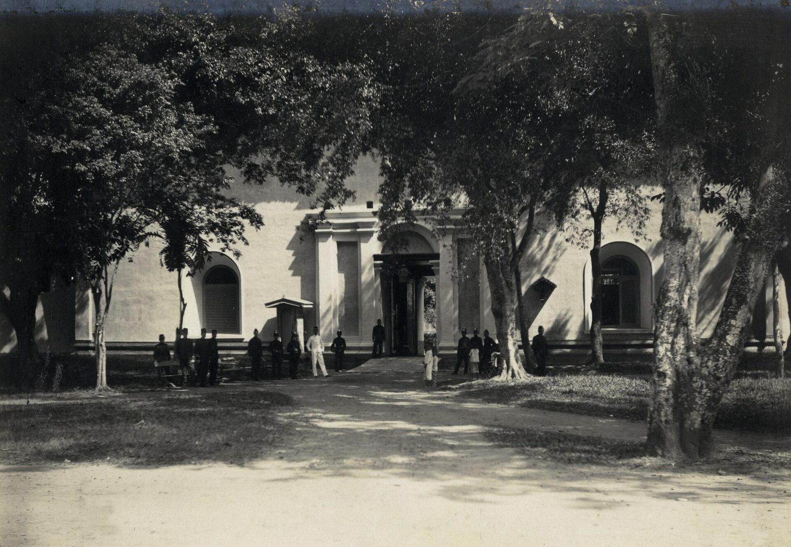 An old photograph of a long whitewashed wall with a arched gateway at its centre, large trees shading the open ground in front of it and a group of small figures standing by the entrance.