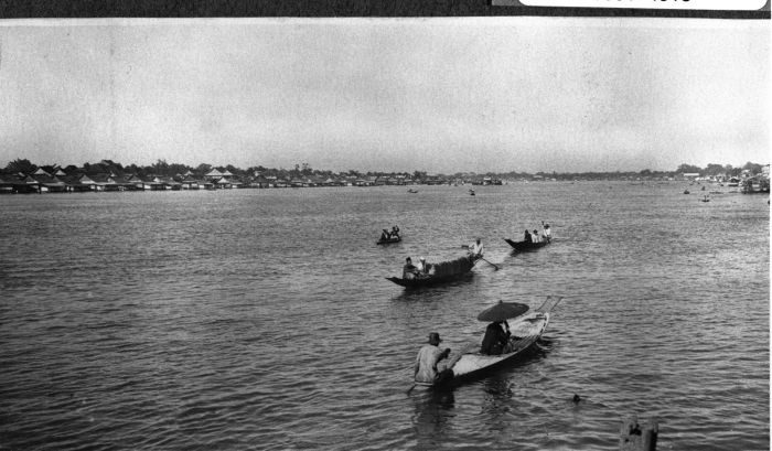 A wide river under a pale sky, two long low boats with awnings being paddled across the middle distance, and a low line of houses along the far bank.