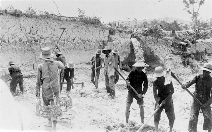 A line of men in wide hats working with long-handled hoes in a shallow open pit, bare earth and spoil banks around them, a few carrying baskets.