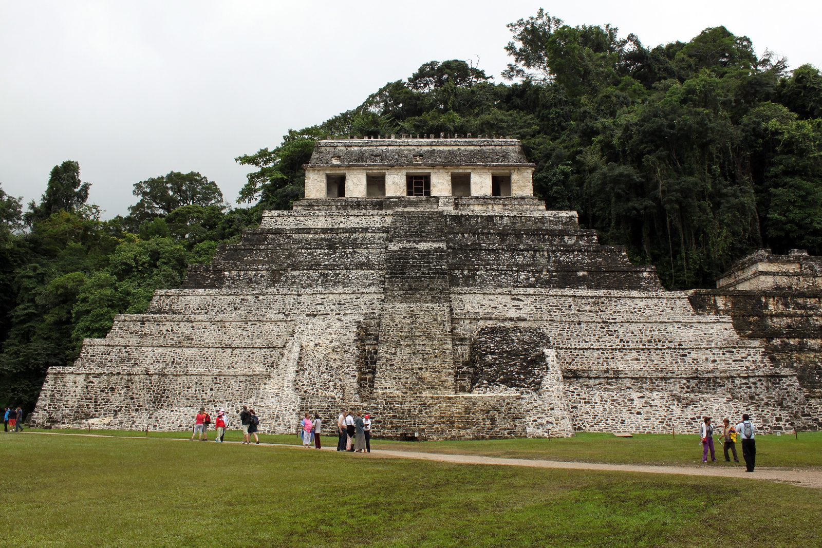 A stepped pyramid with a temple on top and a wide central stair, visitors on the lawn in front.