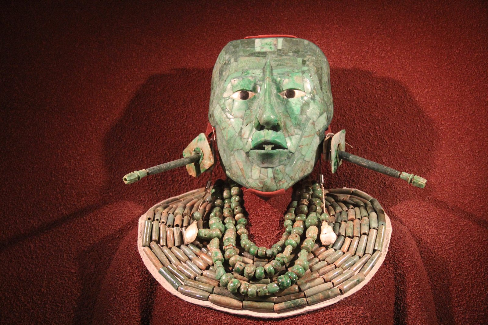 A jade mosaic face mask with shell and obsidian eyes above a broad bead collar, on red cloth.