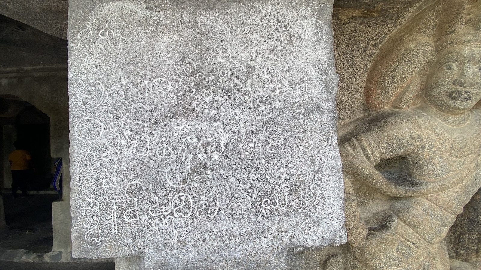 A carved granite pillar face covered with lines of Grantha script beside a guardian figure.