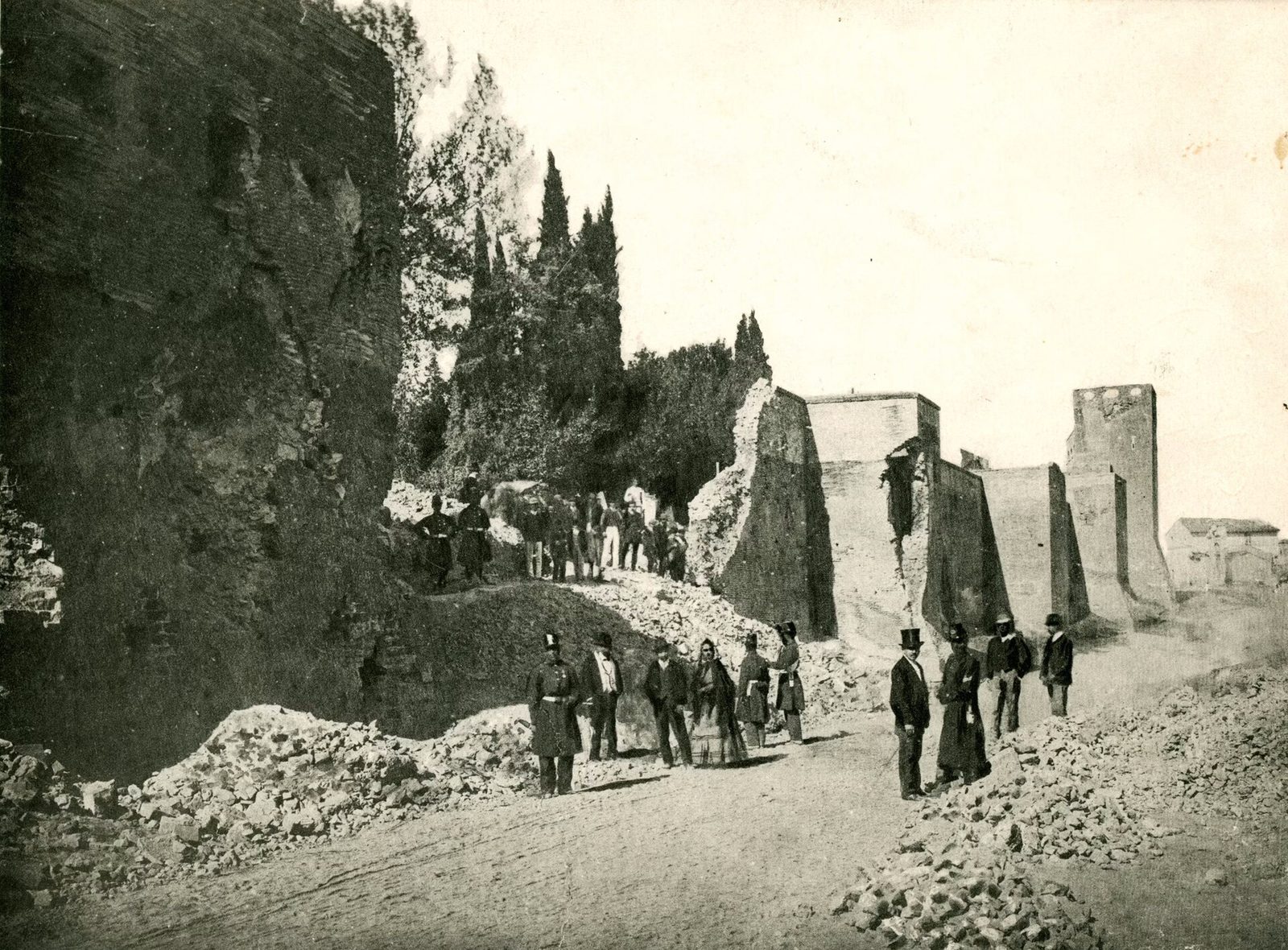 An early photograph of a city wall with a wide ragged breach torn through it, rubble heaped at the foot and a gate structure standing to one side.