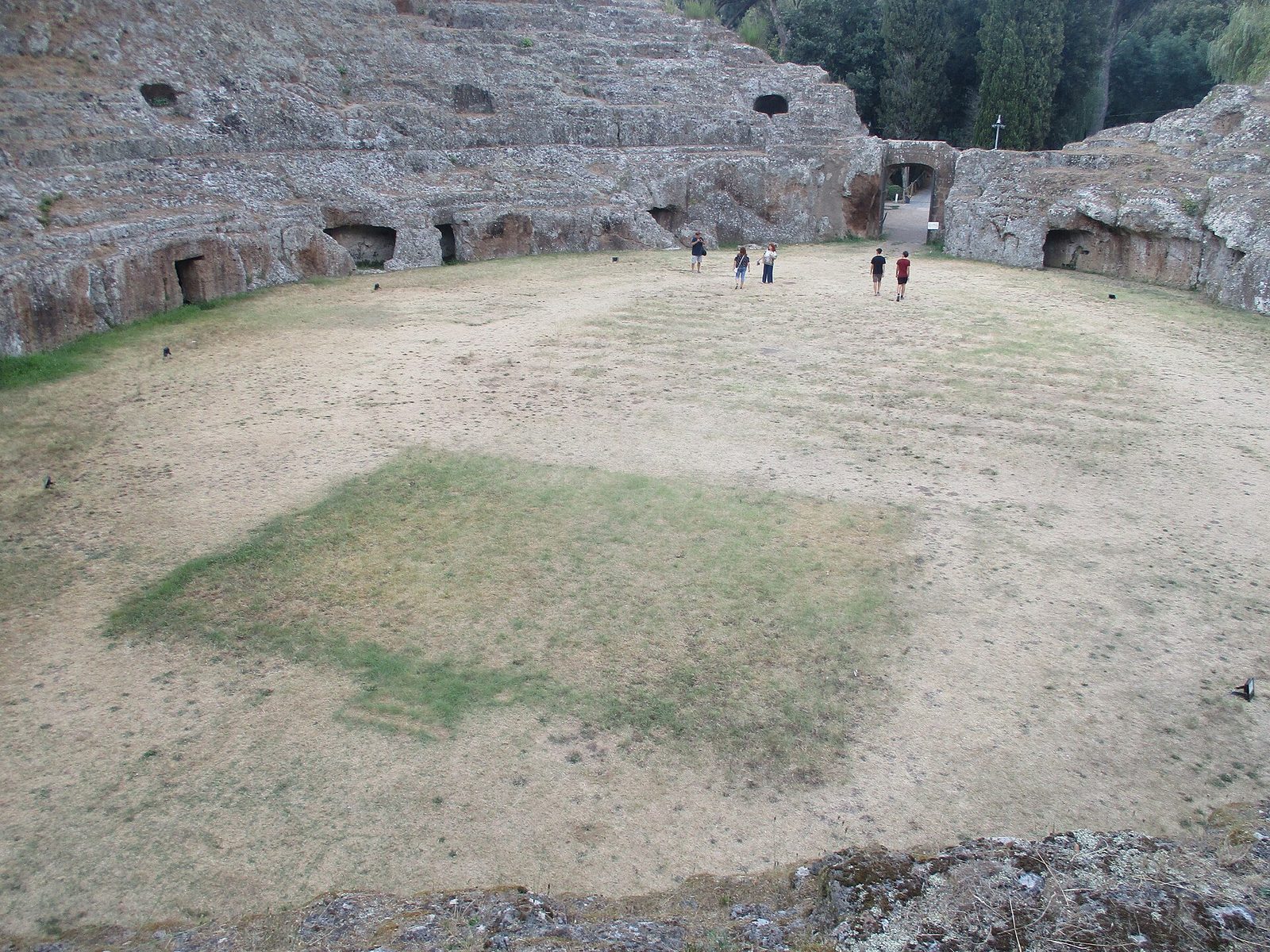 An oval amphitheatre cut down into living rock, its tiers of seating and arched entrances carved out of the stone, surrounded by trees.