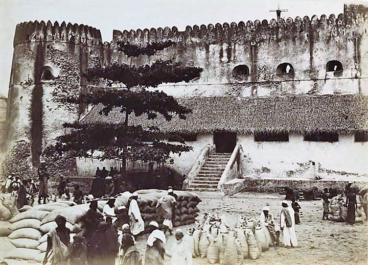 An old photograph of a crenellated fort with a stair to its entrance, a crowd and sacks of goods in the square before it.