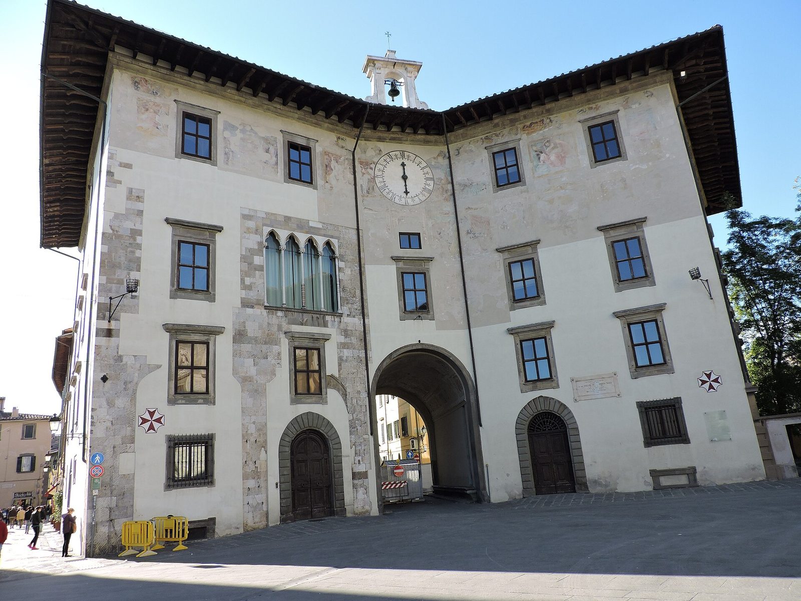 A palace front with a large clock face in its upper gable, joined to its neighbour by an arch over a street, facing an open square.