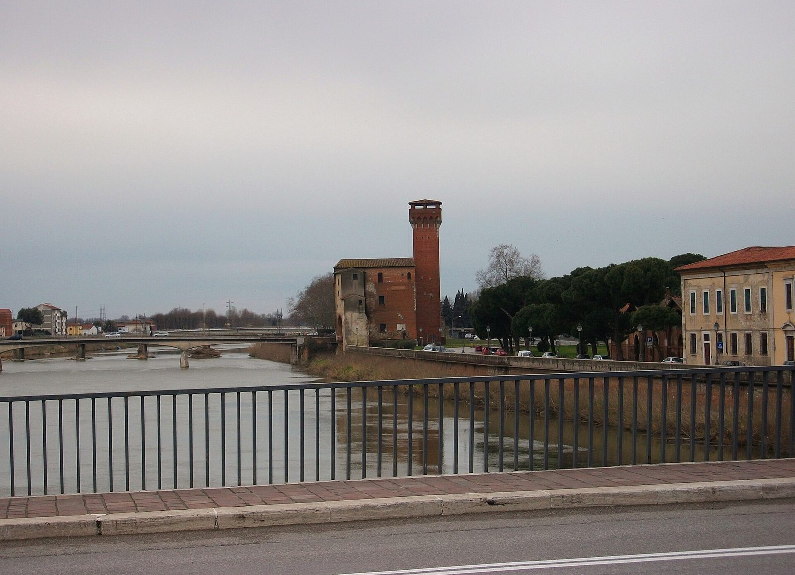 A squat crenellated brick tower at the end of a fortified wall beside a river, with a modern road and trees in front of it.