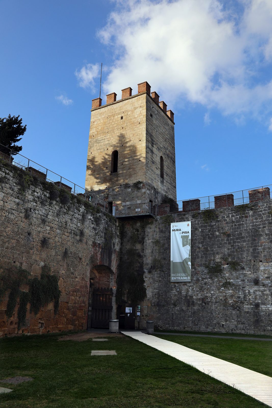 A square brick tower rising from a long run of medieval city wall, with grass at its foot and a clear sky behind.
