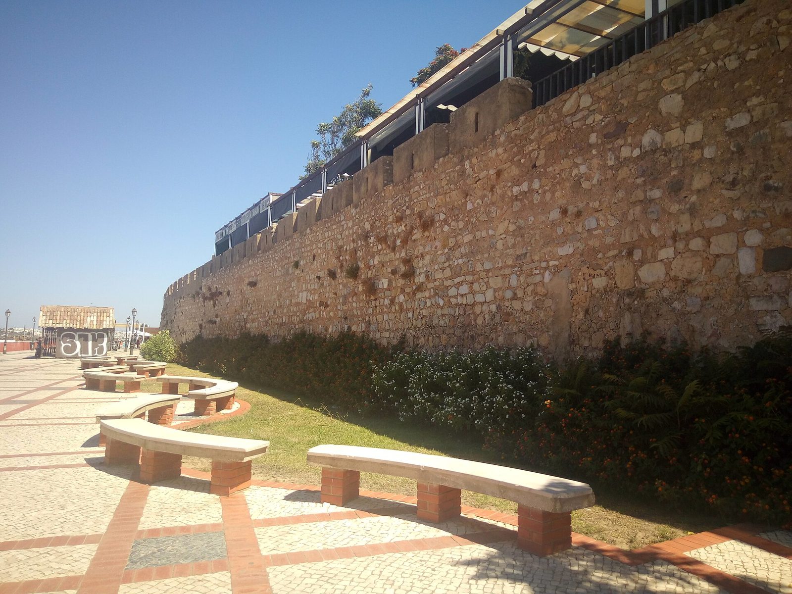 A long stretch of old stone town wall above a paved promenade with benches.