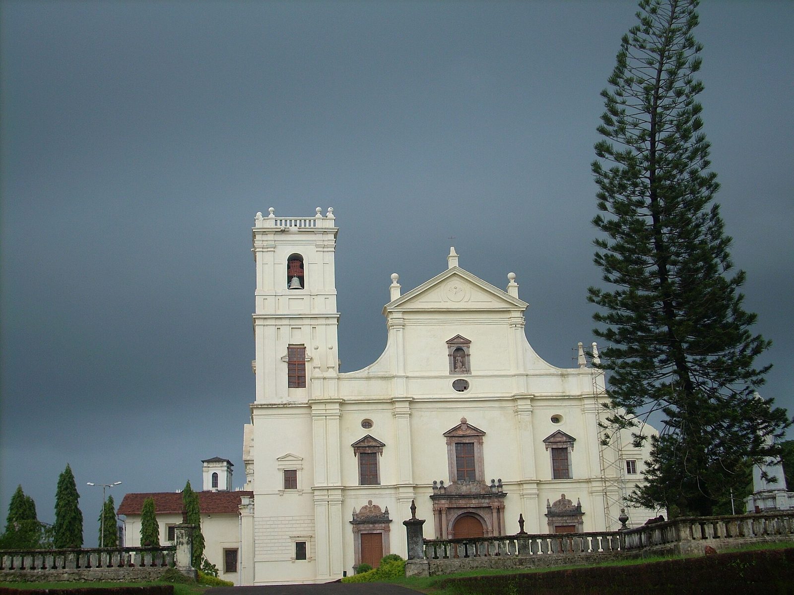 A large white church front with a square bell tower under a dark sky, a tall pine beside it.