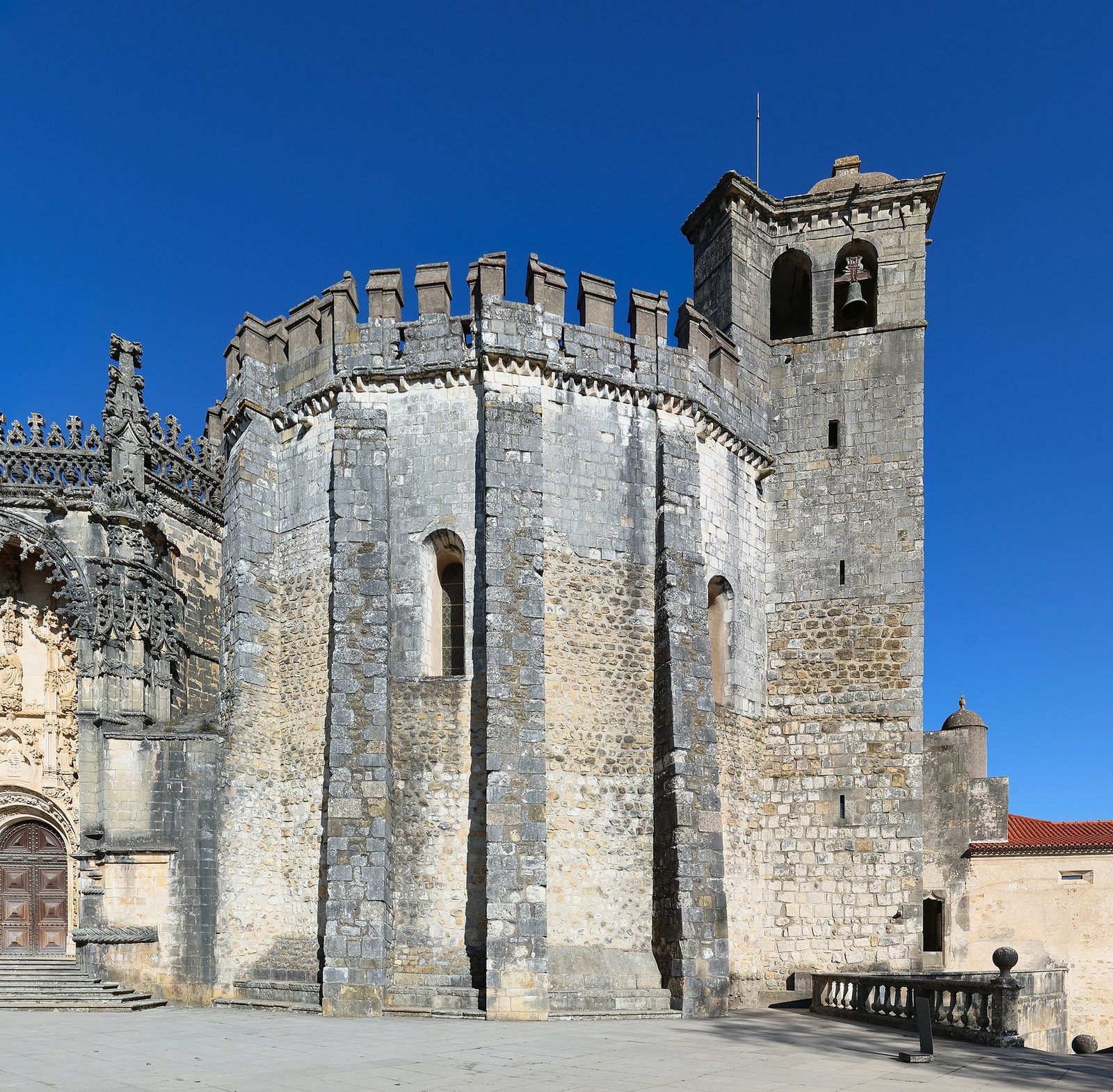 A many-sided Romanesque church of grey stone with a crenellated parapet and a bell tower, under a blue sky.