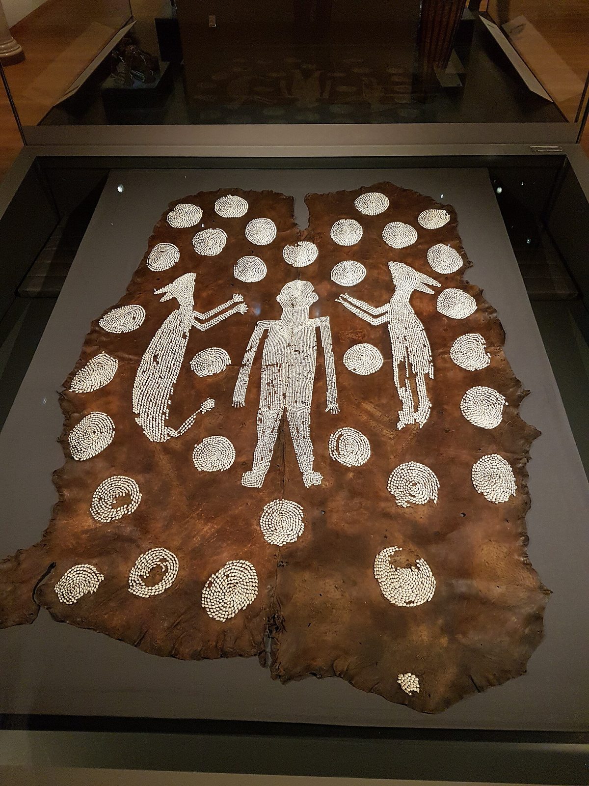 A large hide cloak sewn with shell beads in the shapes of a standing man, two animals and many small circles, in a museum case.