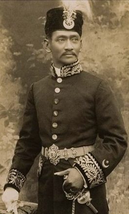 A sepia photographic portrait of a man standing in a dark high-collared uniform with rows of buttons and heavy braid, a crested cap on his head and one hand resting on an ornamented dagger at his waist.