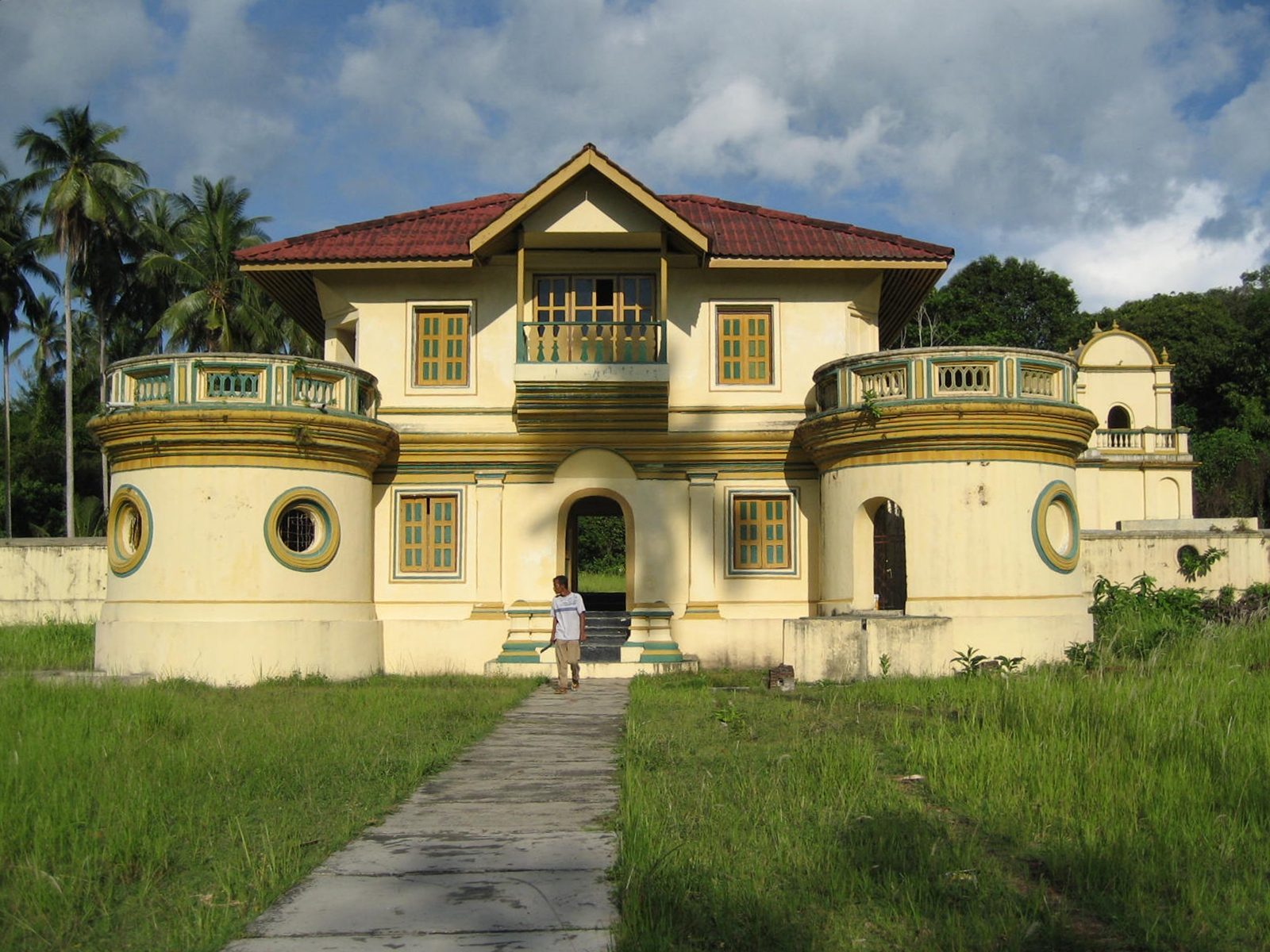 A two-storey stuccoed building with a red tiled roof and a shuttered upper balcony, its ground floor flanked by round turret-like bays with porthole windows, standing empty behind a rough path through long grass.