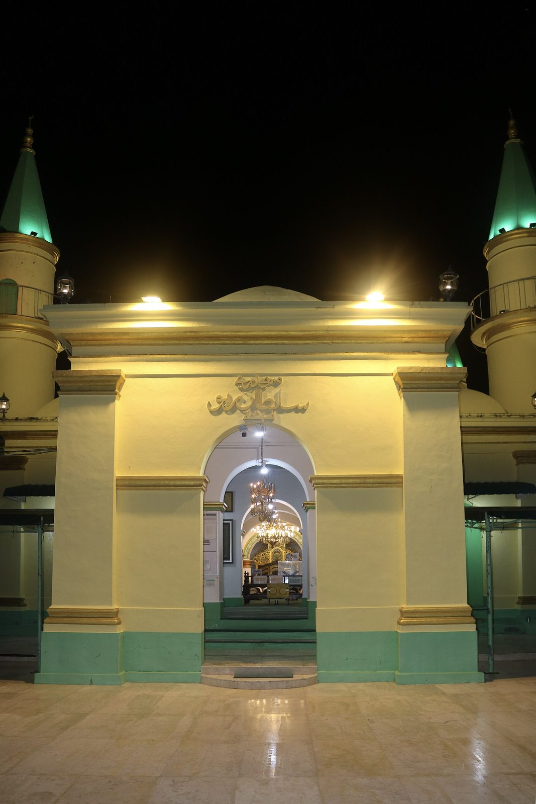 A mosque gateway lit from below at night, yellow walls with green trim, an inscribed panel over the arch and chandeliers visible through the opening, with green-capped minarets rising on either side.