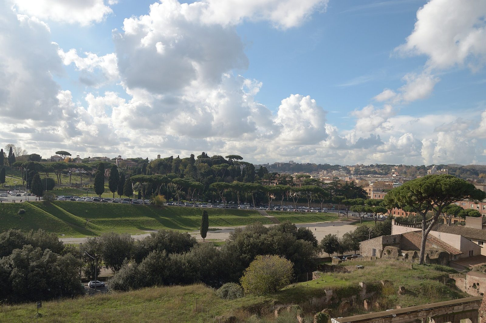 A long flat grassed valley between two hills, its oval outline still legible in the ground, with a low bank running down one side and the roofs of a city beyond.