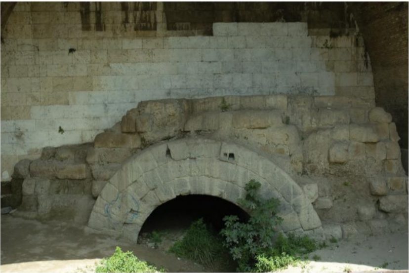 A stone-faced river embankment pierced by a large arched opening at water level, the voussoirs of the arch set into regular ashlar blocks.