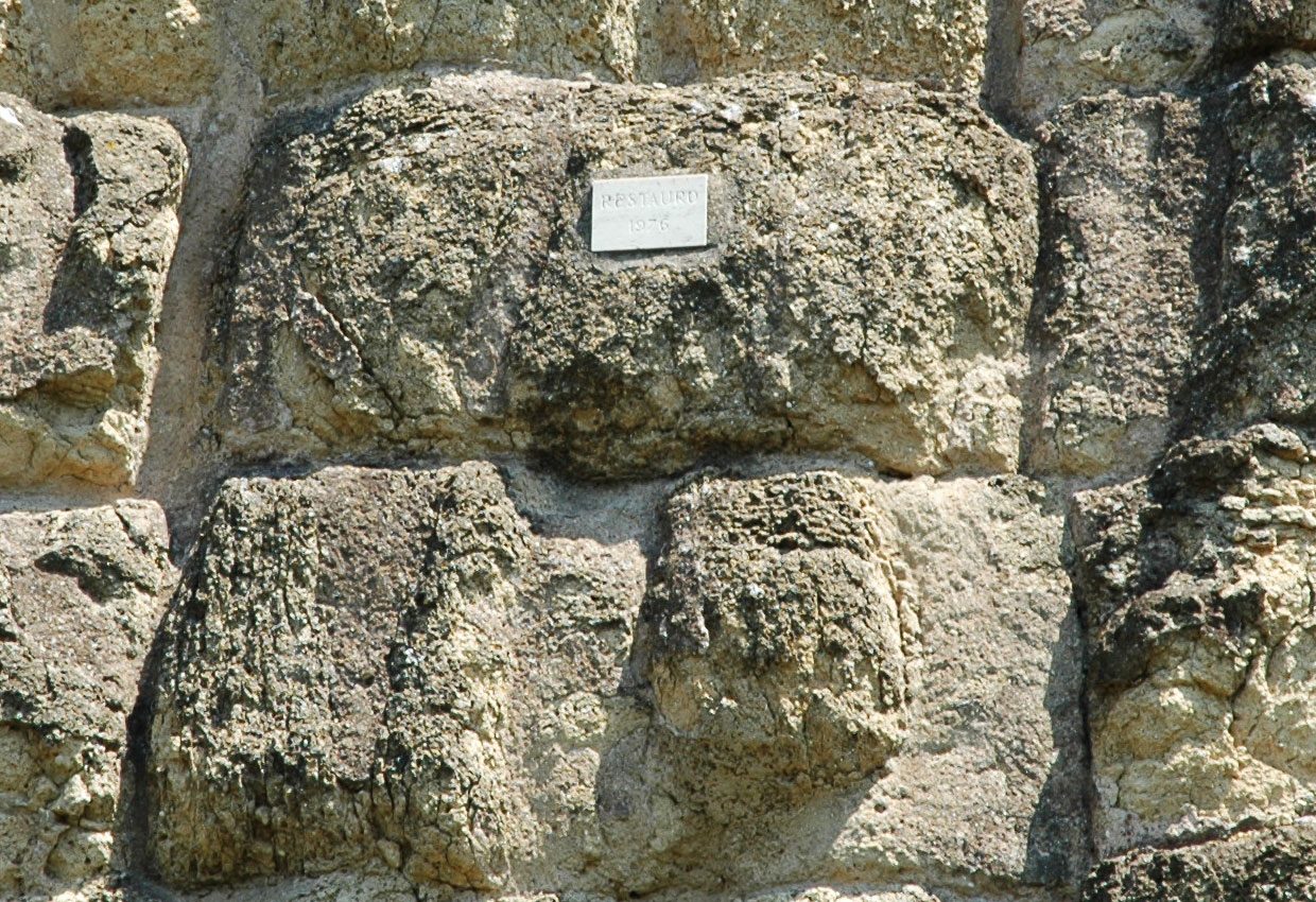 A close view of the wall's stonework: large blocks of pale grey-yellow volcanic rock, deeply pitted and pocked across their faces, with a small printed museum label fixed to one of them.