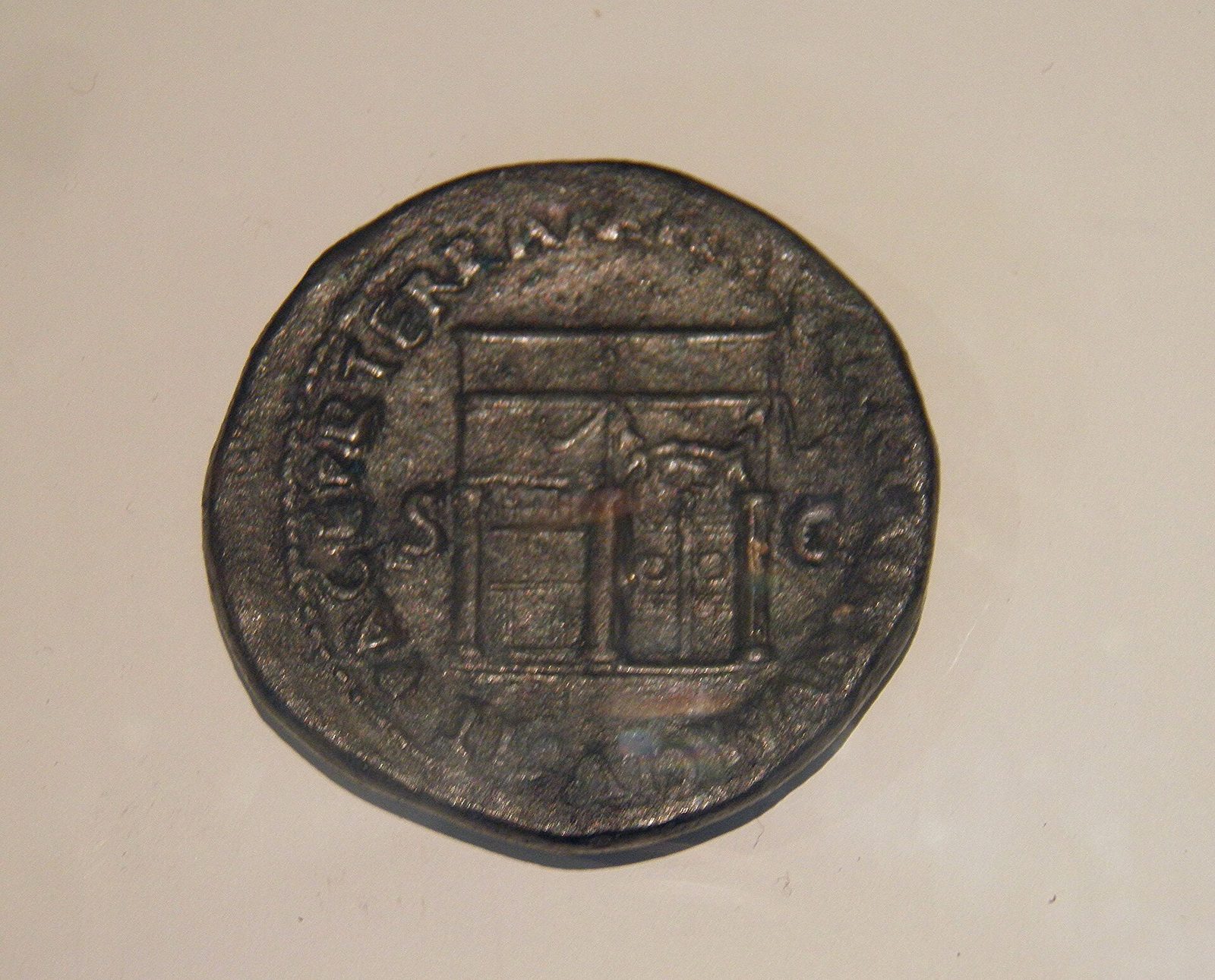 A large worn brass coin showing a small temple building in side view, its double doors shut and garlanded, with a Latin legend running round the rim.