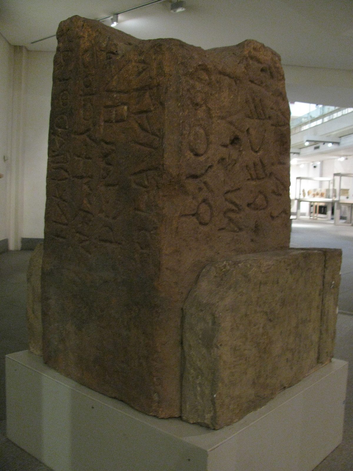 A broken block of tufa standing on a dark floor, its faces covered in columns of archaic letters cut vertically, the top of the stone sheared away.