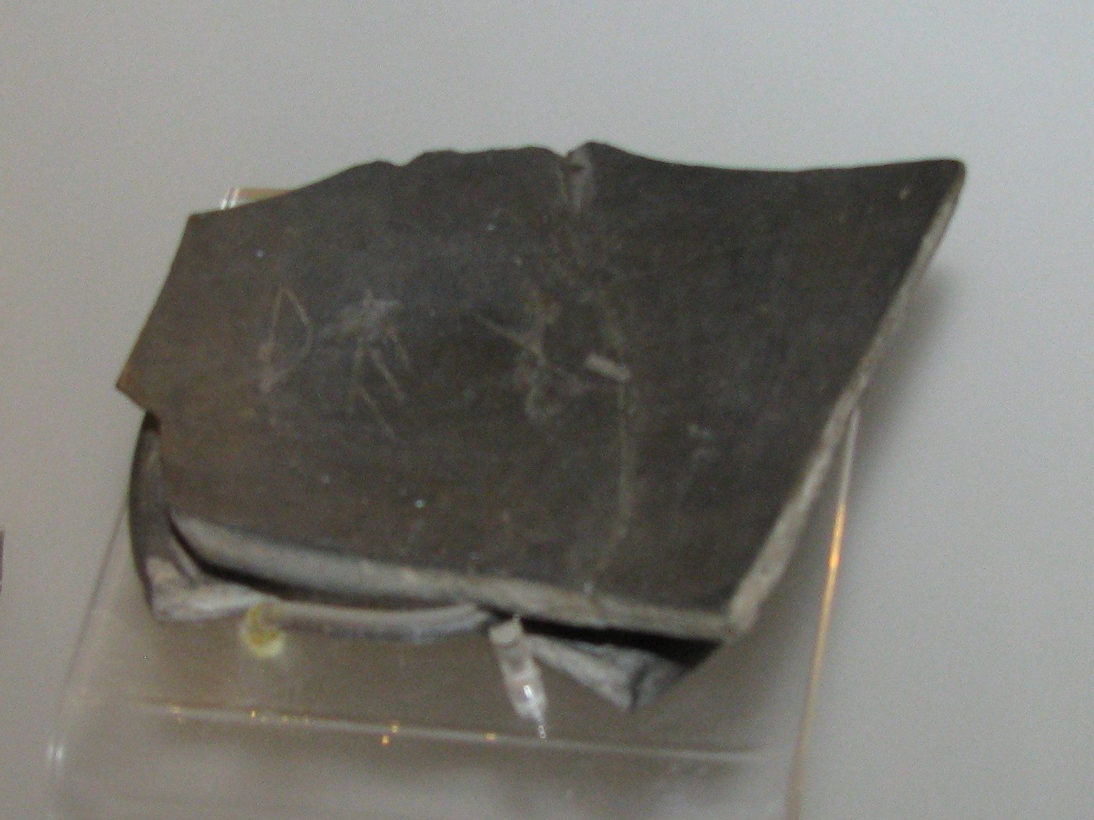A fragment of black pottery with a sheared edge, displayed on a mount against a pale wall, a few faint scratched strokes just visible across its dark outer face.