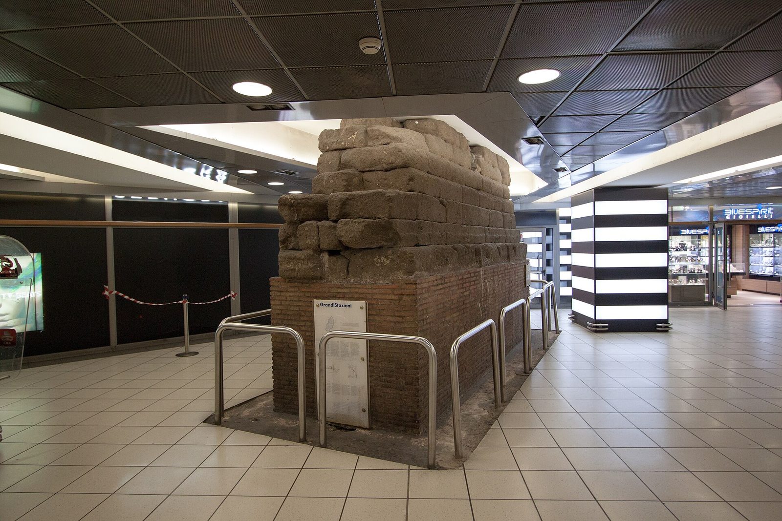 A high wall of large rectangular stone blocks laid in regular courses, standing incongruously inside a modern concourse of glass and polished floor.