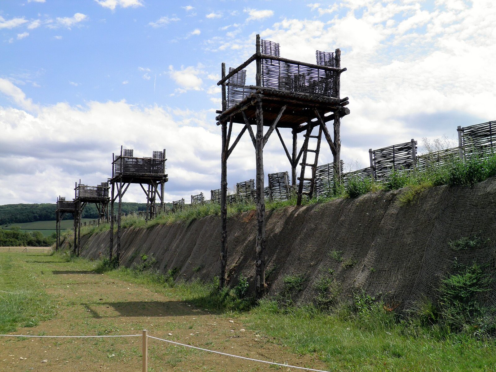 A reconstructed timber rampart with a palisade and towers behind a wide ditch, rows of sharpened stakes planted in the open ground in front of it.