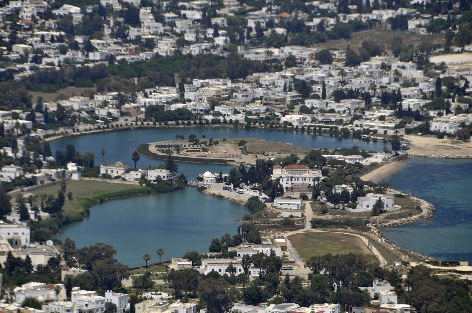 A circular lagoon with a small island at its centre, ringed by grass and low modern houses, with open water and a coastline beyond.