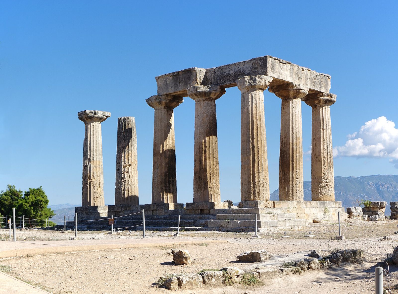 Seven massive fluted limestone columns with heavy capitals standing on a rock platform above a modern village, with a steep mountain rising behind them.