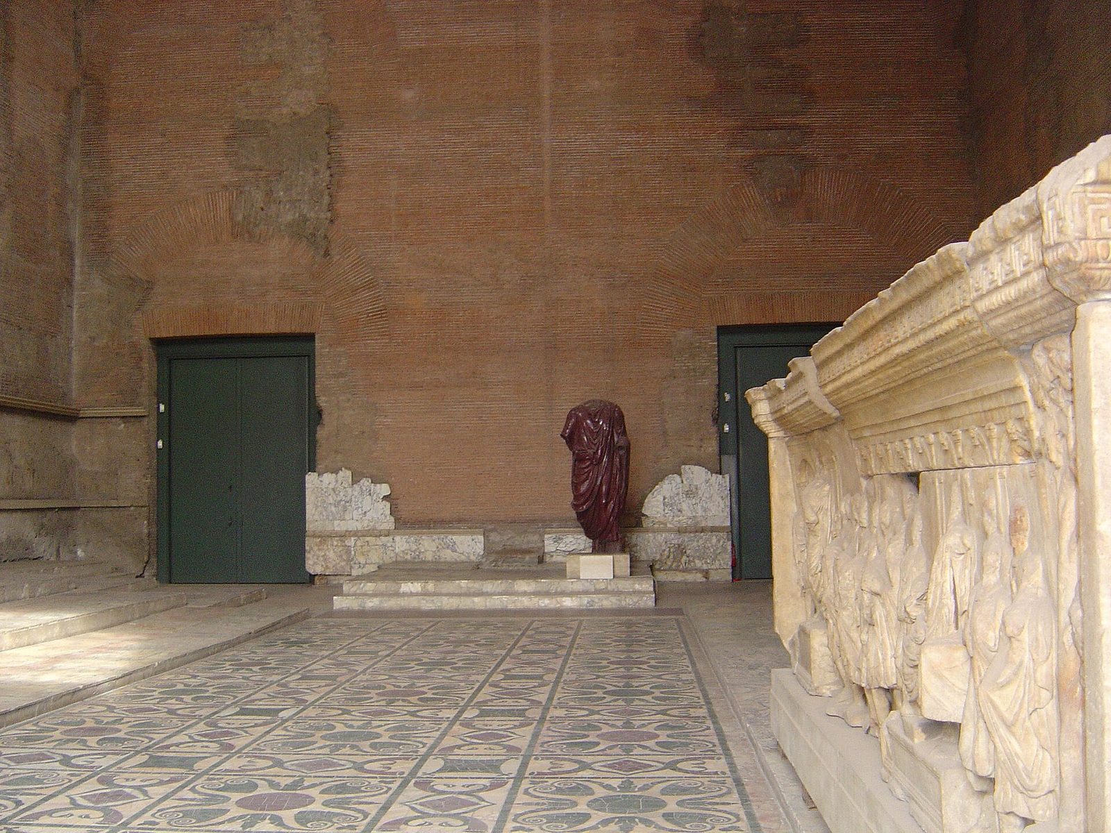 A tall plain hall with a patterned marble floor, broad low steps running along both side walls where seating stood, and a small raised platform at the far end under high windows.