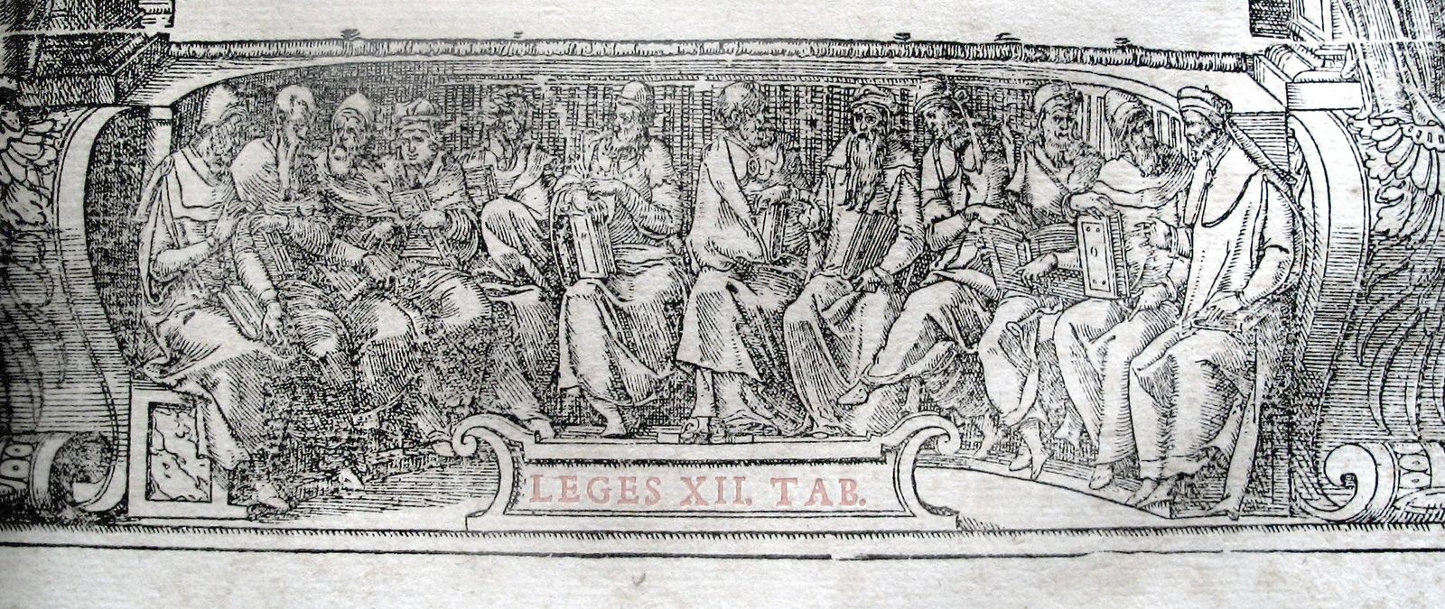 An engraved frieze of robed men seated in a row along a curved bench, several holding hinged writing tablets open on their knees, with the words LEGES XII TAB lettered in red on a cartouche below.