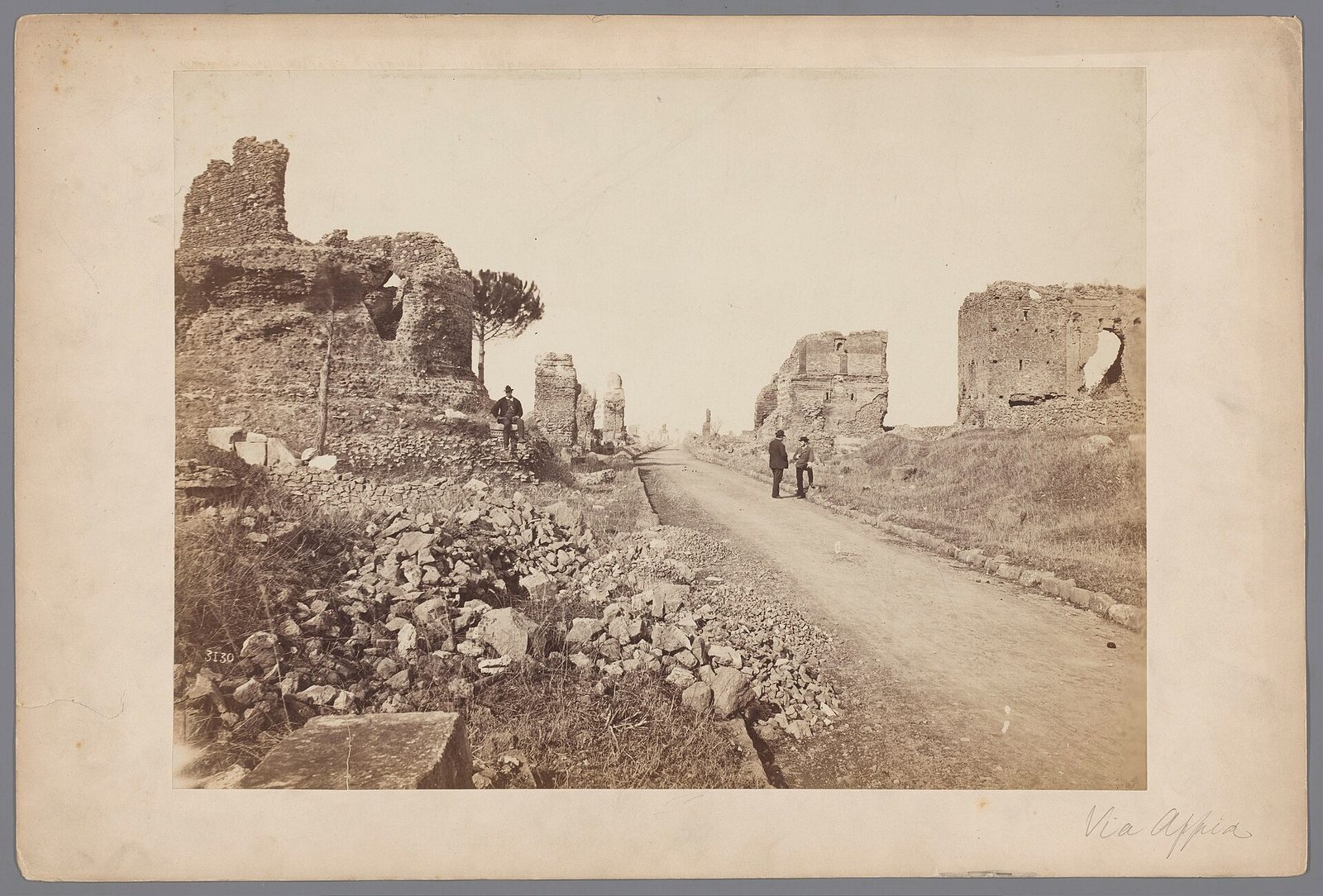 An early photograph of a straight paved road of large irregular basalt slabs running to the horizon between low walls and ruined tombs, with flat country on either side.