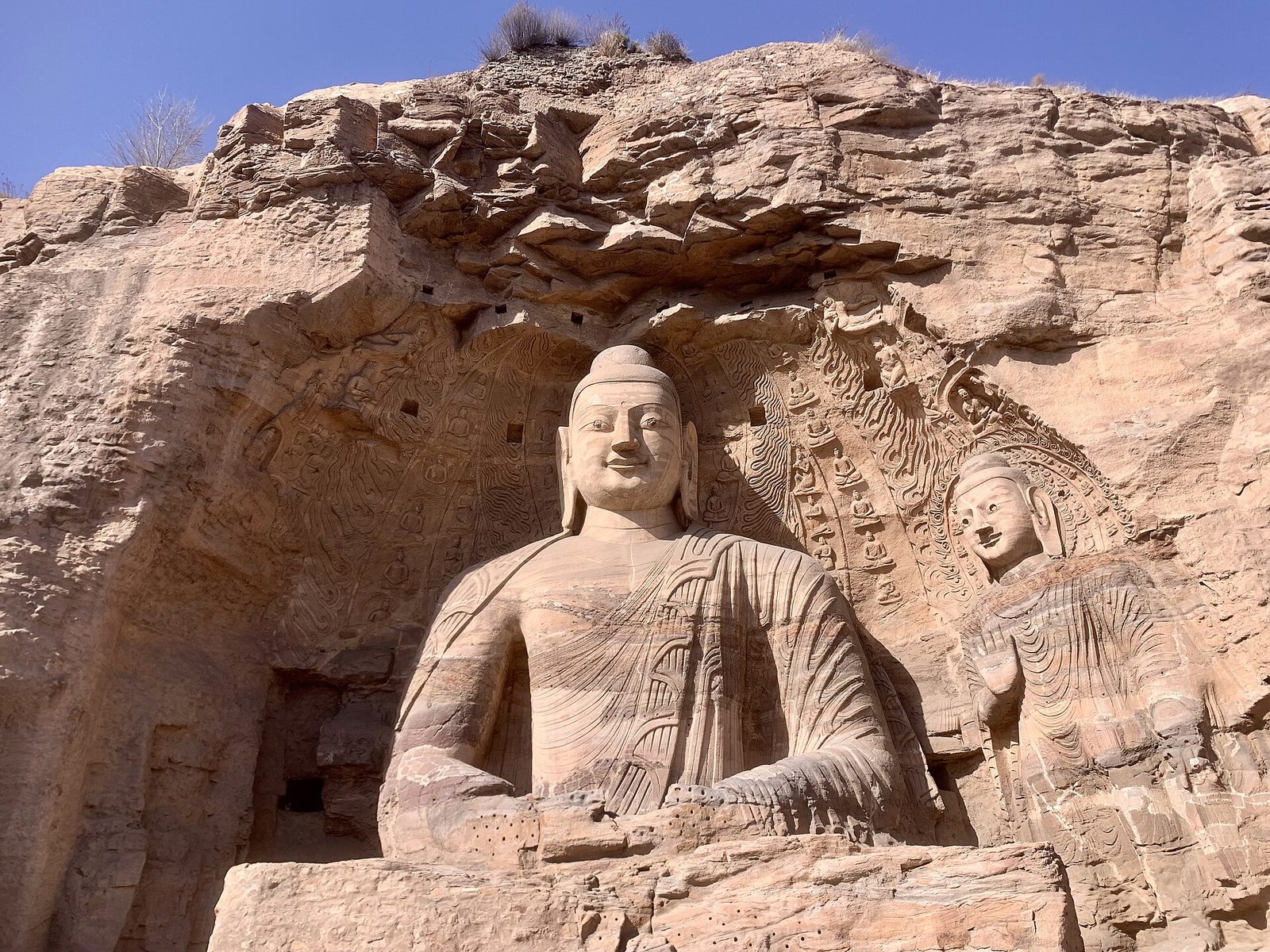 A colossal seated Buddha carved into a cliff face, a standing figure beside it, under an overhang of crumbling rock.