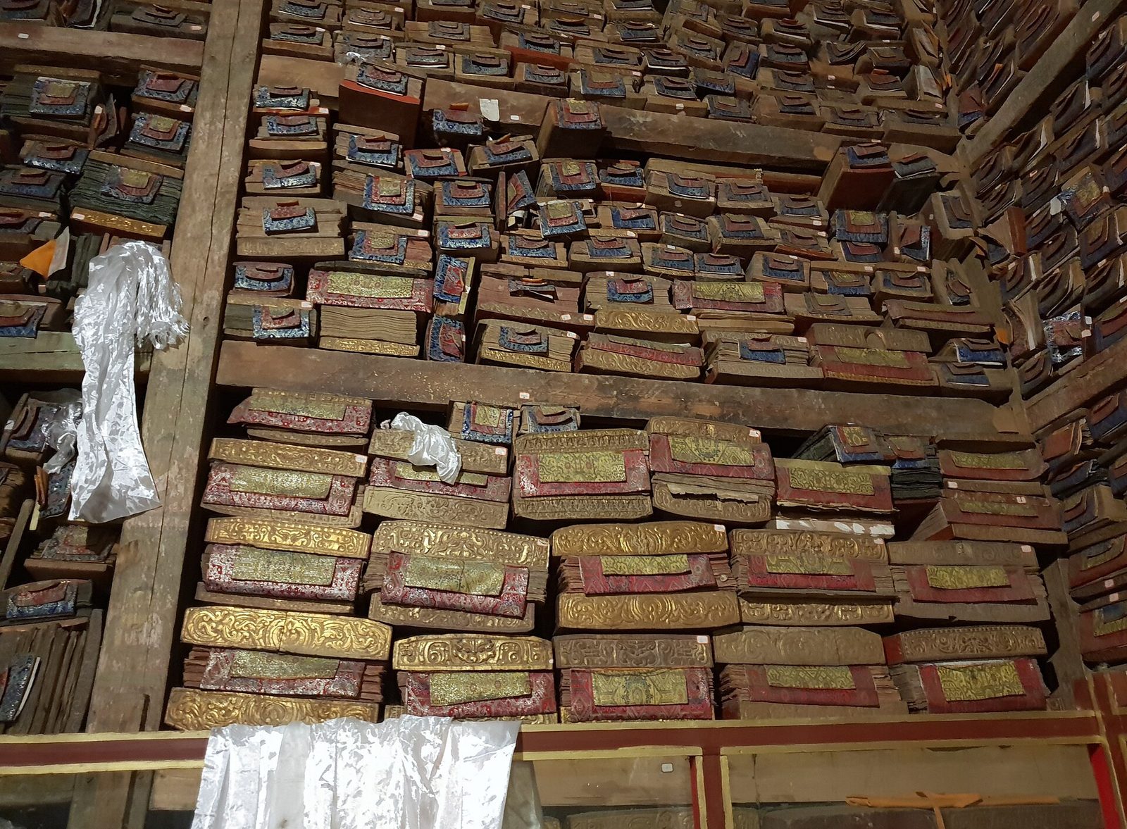 Floor-to-ceiling wooden shelves packed with wrapped Tibetan books, white scarves hanging in front.