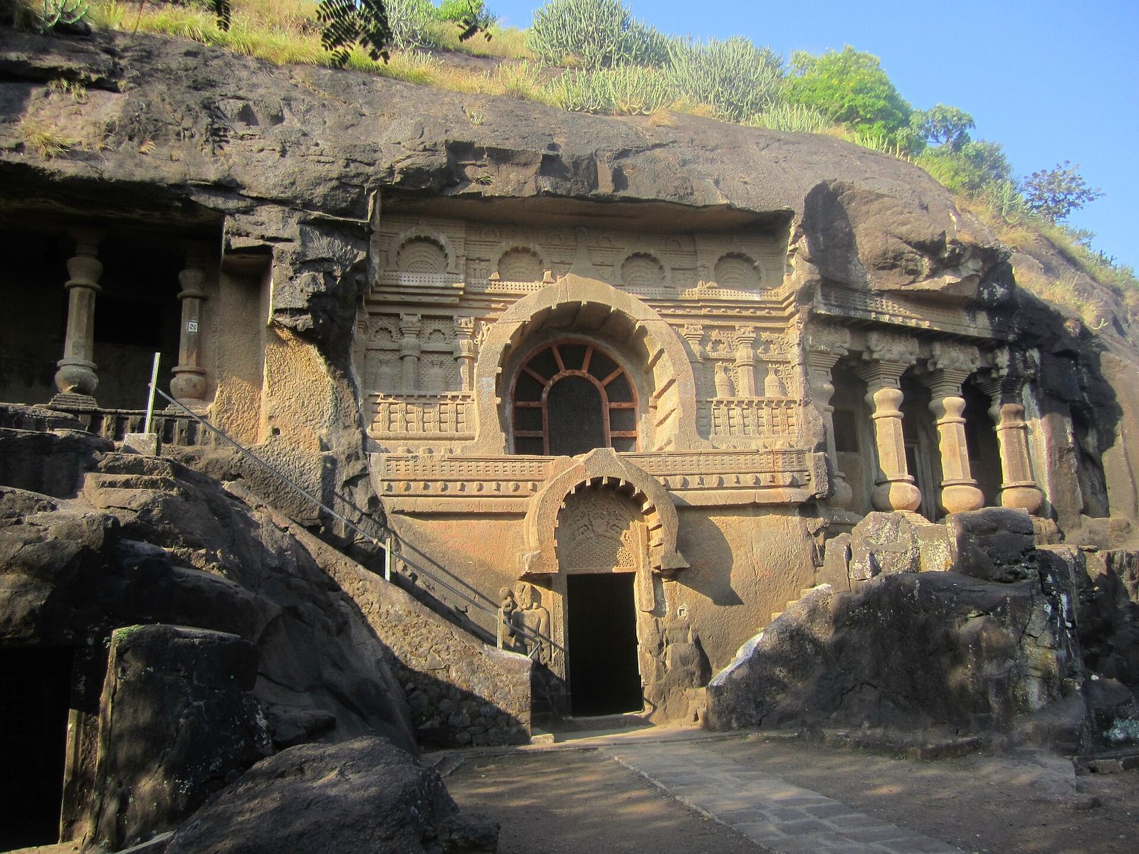 A rock-cut cave front with a horseshoe-arched window and carved pillars in a cliff.