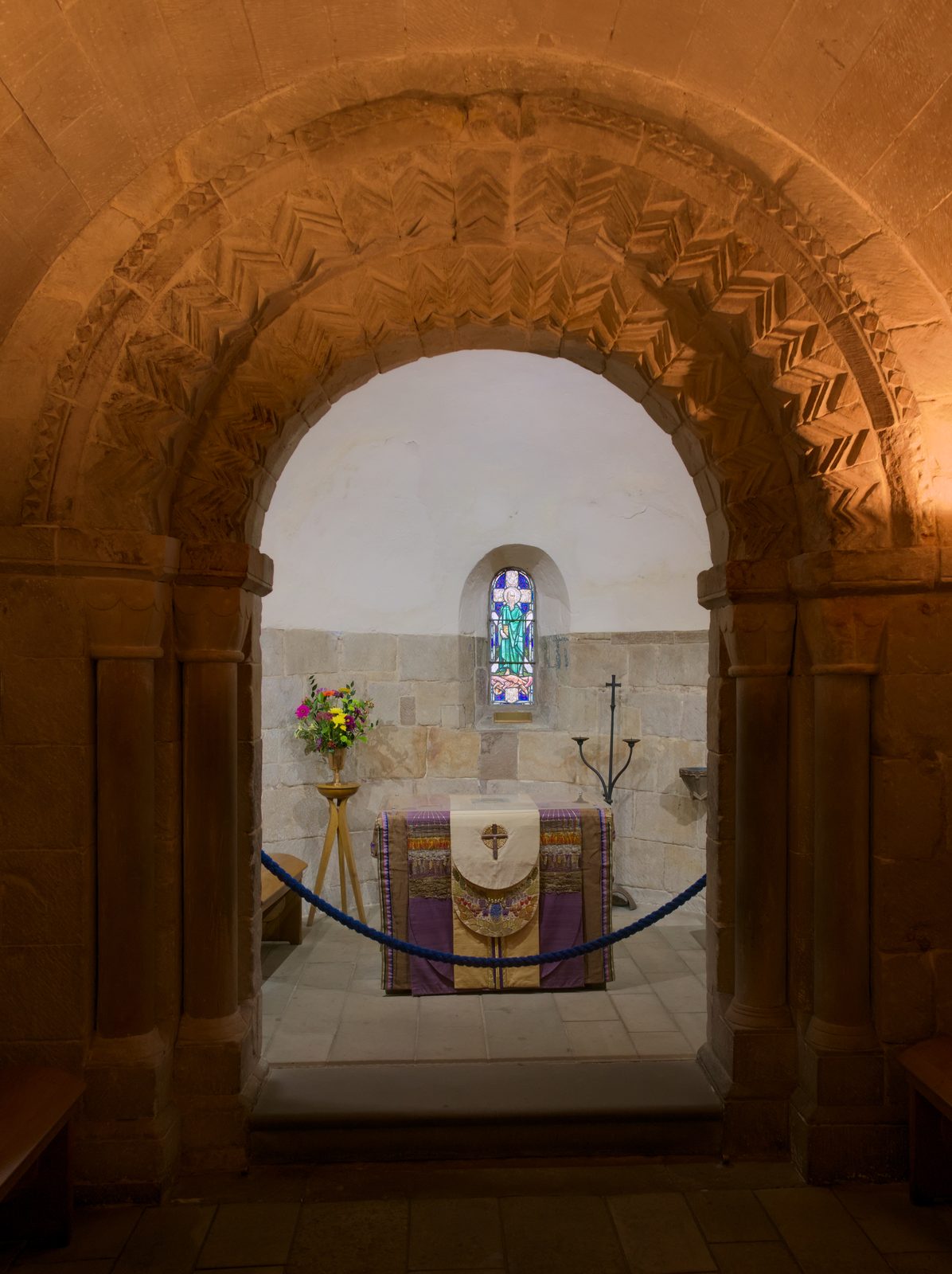 A small stone chapel seen through a round arch carved with zigzag ornament, an altar beyond.