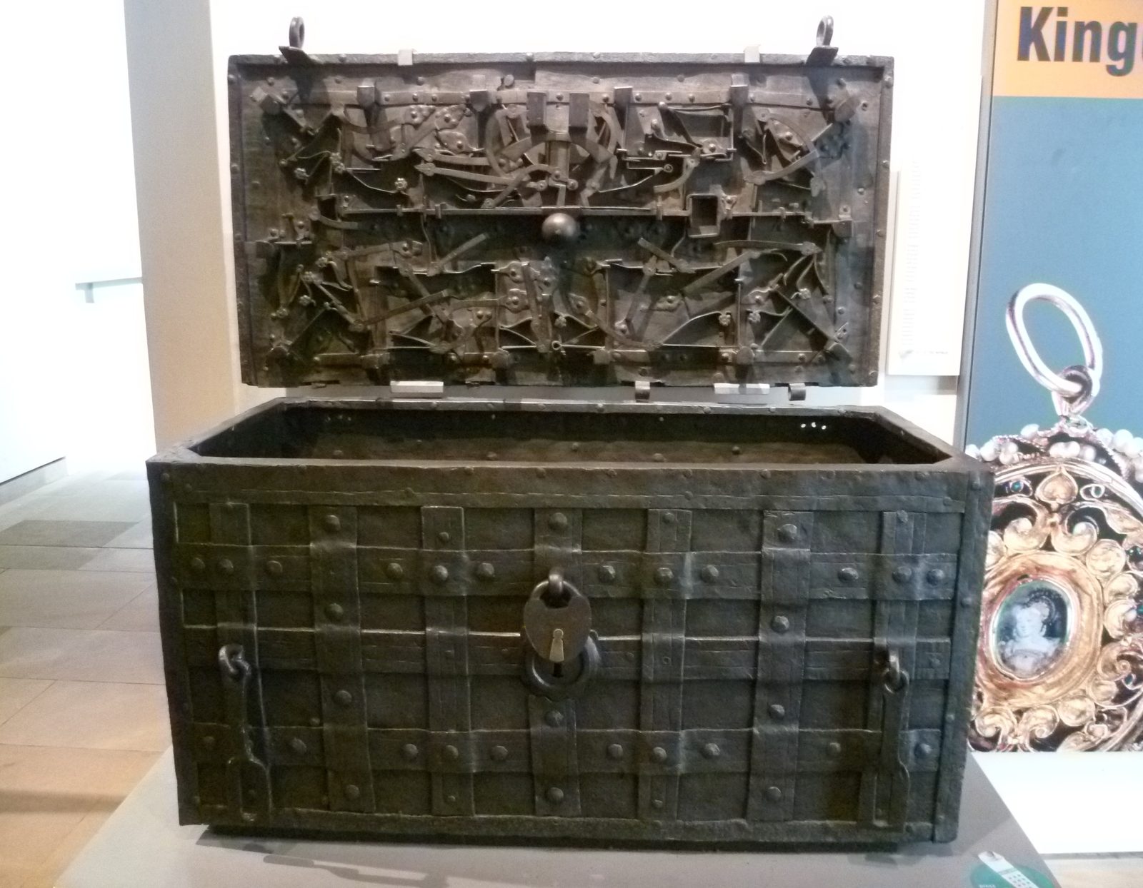 An iron-bound strongbox with an elaborate locking mechanism exposed in its open lid, in a museum case.