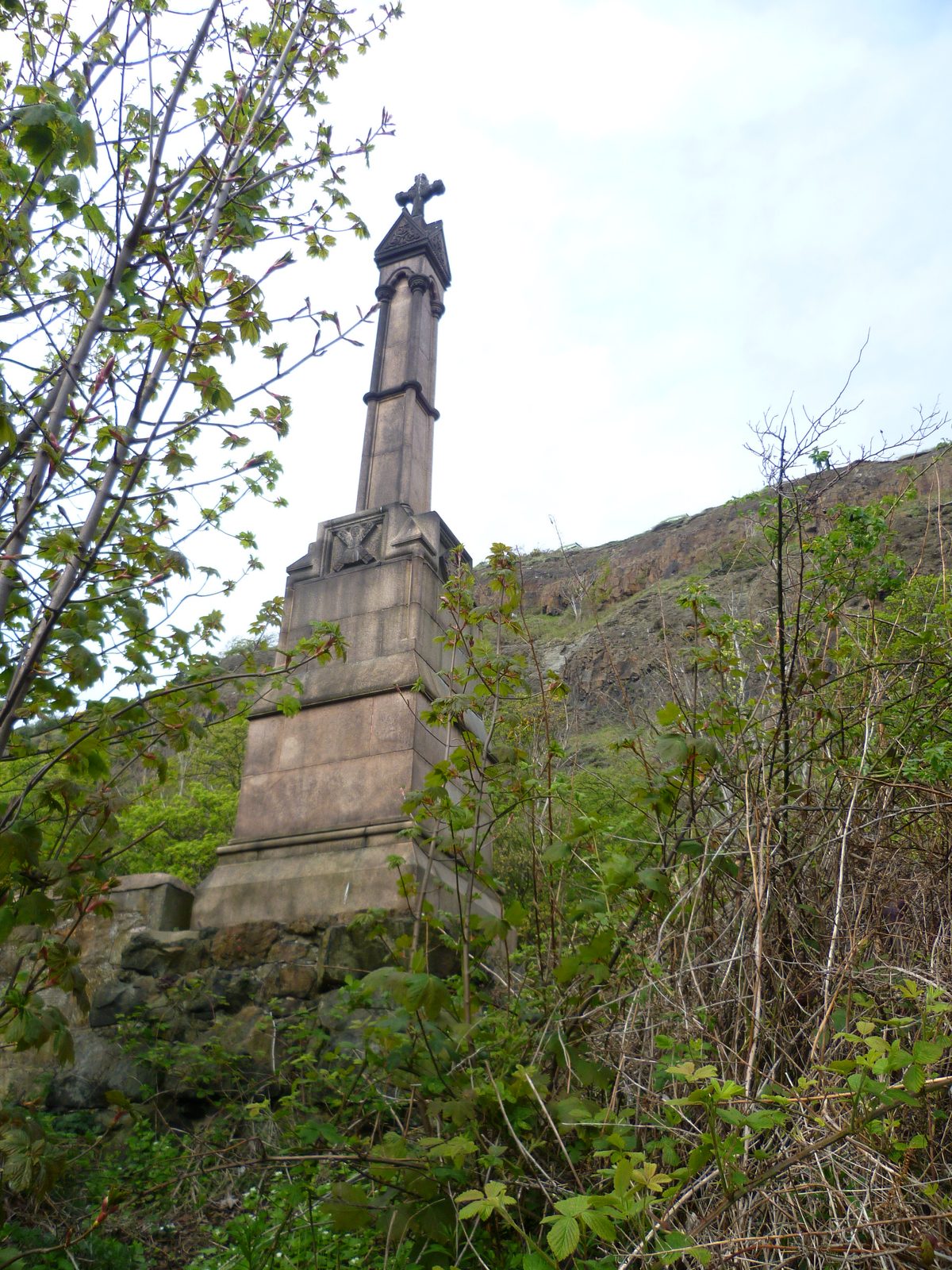 A stone column topped by a cross, standing among trees on a hillside.