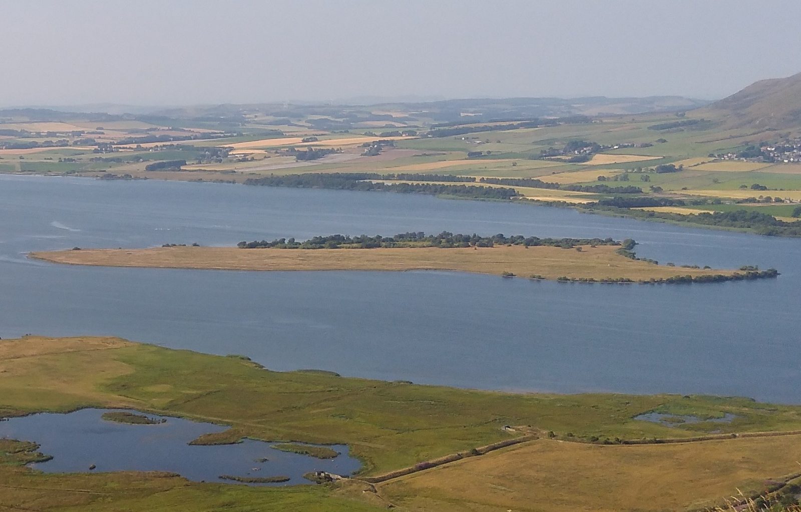 A long low island in a loch, seen from a hillside above farmland.