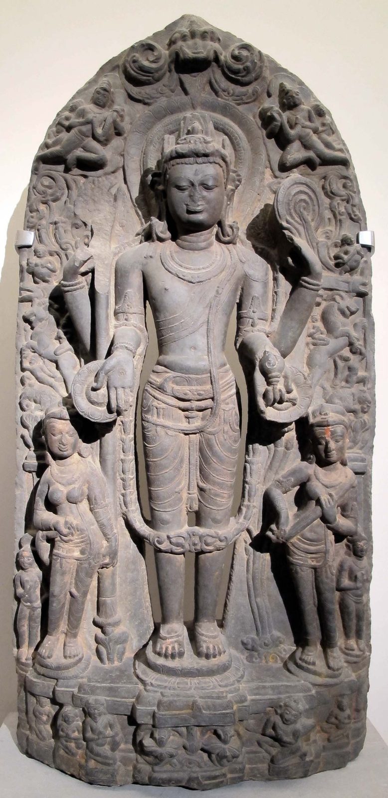 A dark stone stele of a standing four-armed Vishnu with two attendant figures.