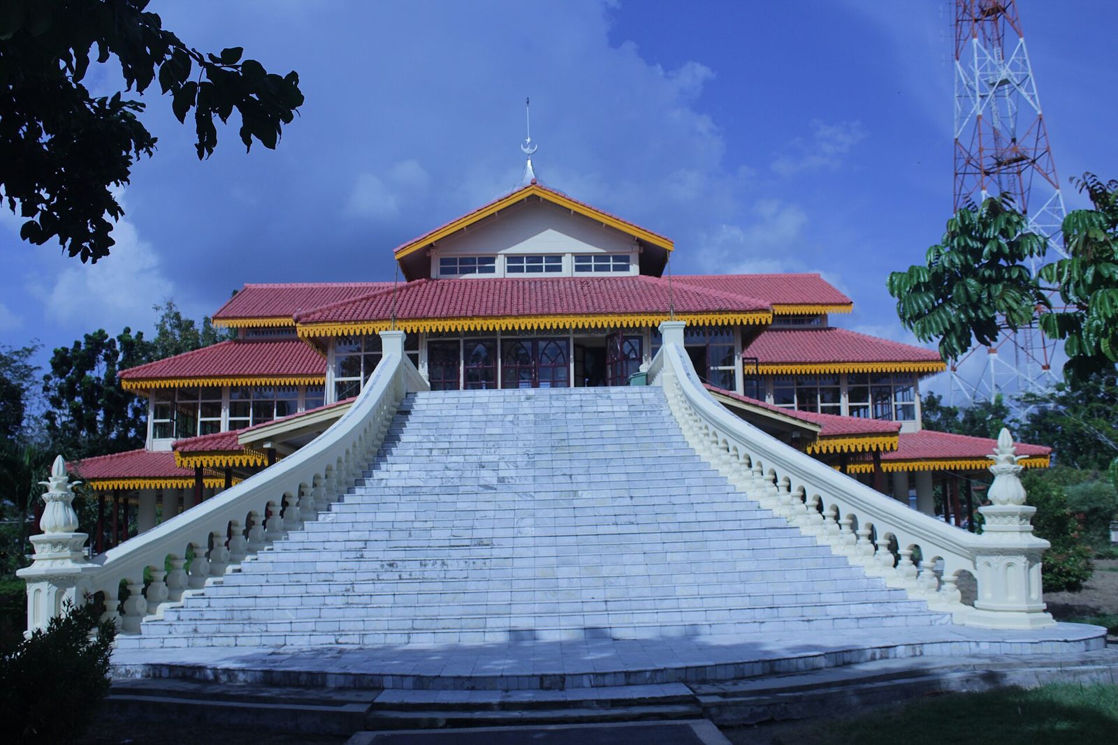 A timber audience hall with a wide tiered red roof and yellow trim, raised on a platform and approached by a broad flight of white steps with balustrades, against a dark blue sky.