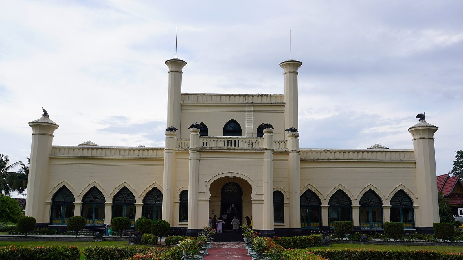 A long cream-coloured palace of two storeys with arcaded arches along the ground floor, square towers at each corner topped with small domes, and clipped hedges and lawns in front.