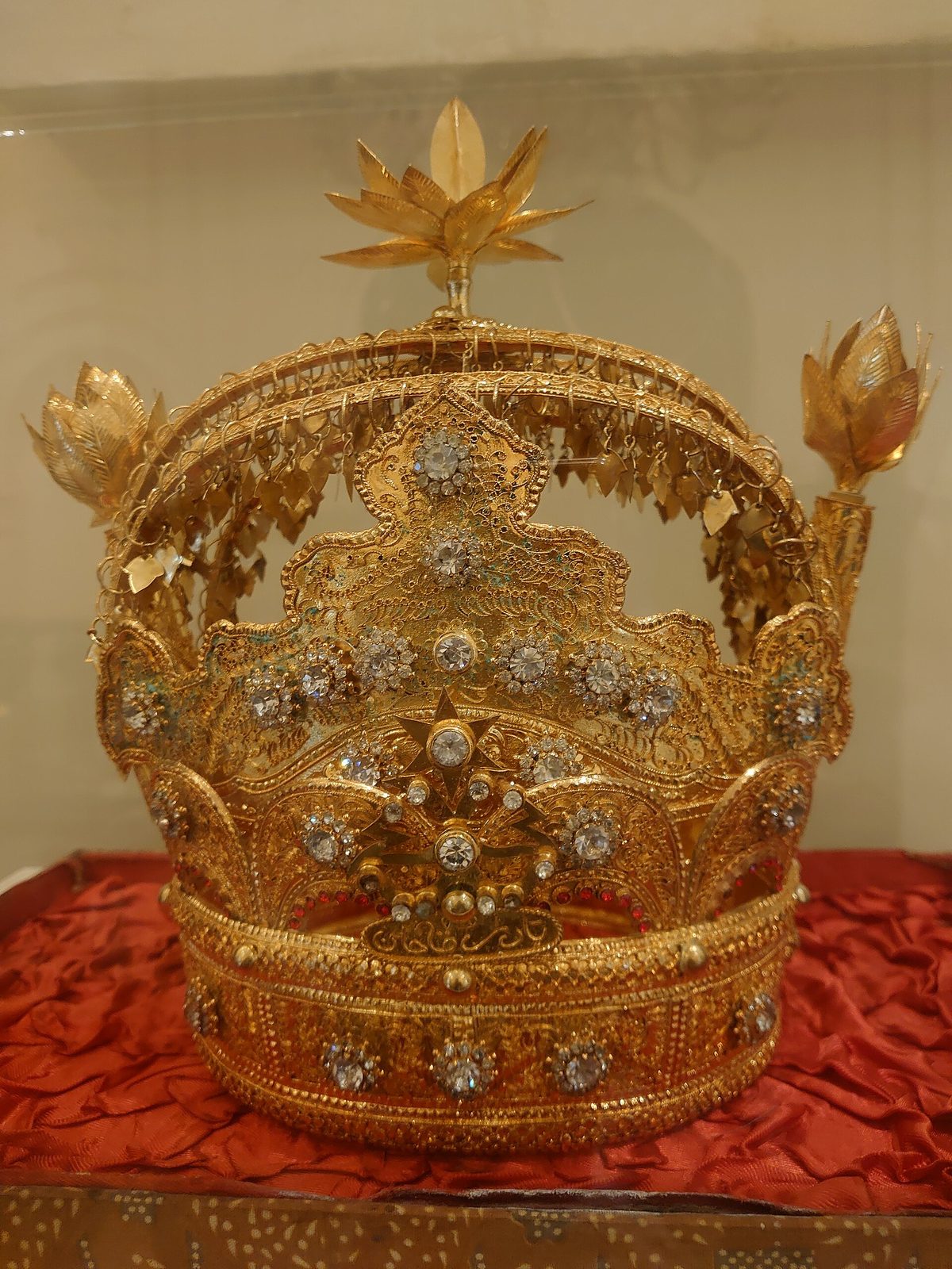 A tall open gold crown in a display case: a jewelled band below, pierced scrollwork above it studded with clear stones, and three leaf-shaped finials rising from the rim.