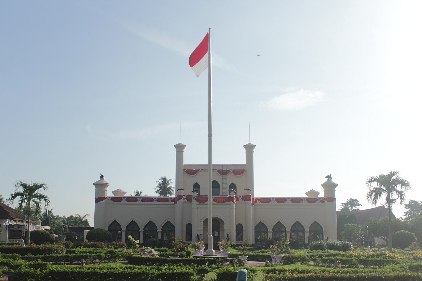 The same cream palace seen across its gardens, with a tall flagpole in front of it carrying a red and white flag against a bright sky.