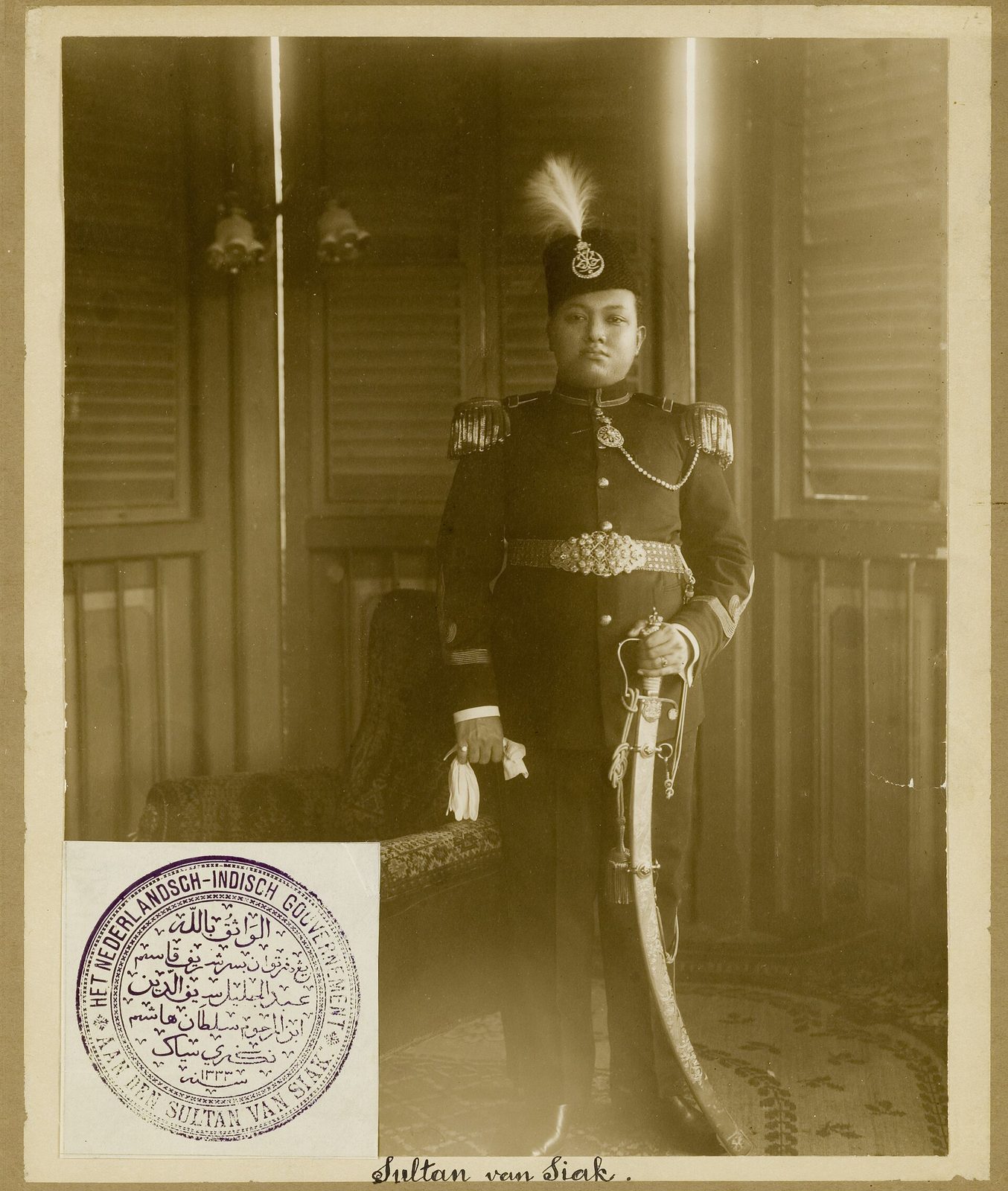 A sepia studio photograph of a young man standing in a dark military tunic with braid, a sash and a plumed cap, one hand resting on a sheathed sword; a stamped seal and a handwritten caption sit in the lower corner of the mount.