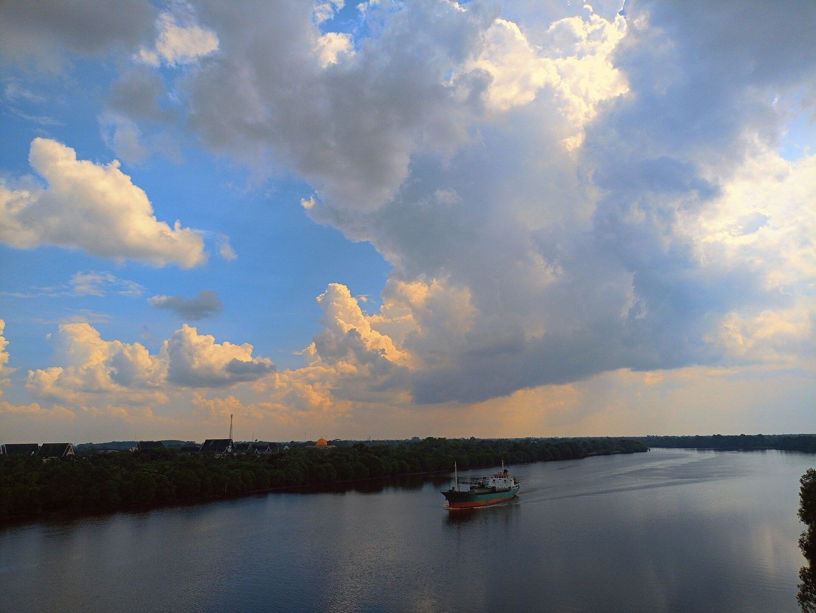 A broad river at evening under a tall sky of broken cloud, low wooded banks on both sides and a single small vessel out in midstream.